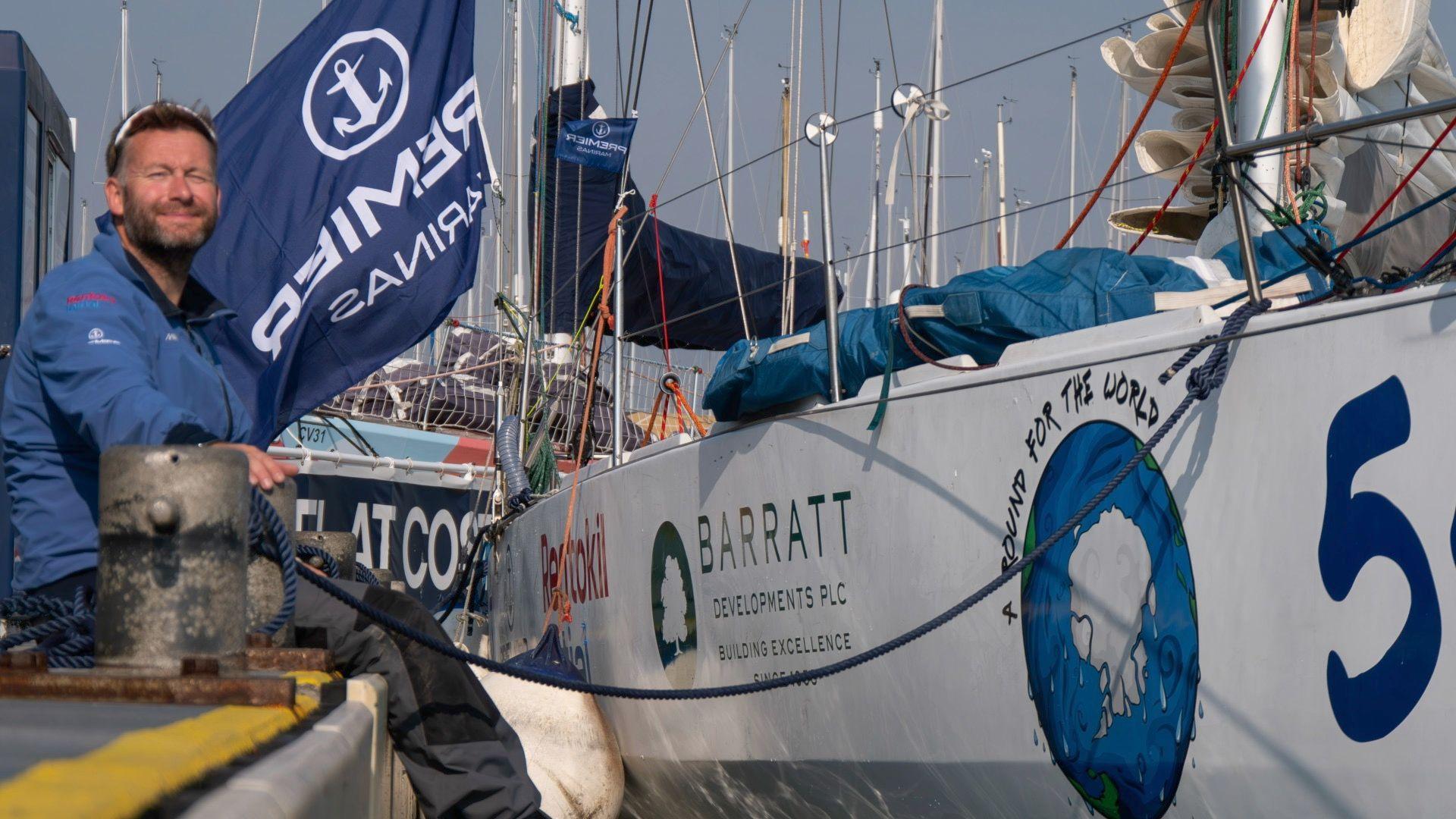 Mr Ketchell is sat on the decking of a marina next to his yacht. He is smiling looking towards the camera and slightly squinting as the sun is on his face. His sunglasses are on top of his head, he is wearing a blue sailing jacket and grey and black cargo trousers. His yacht is white and covered in business sponshorship logos.