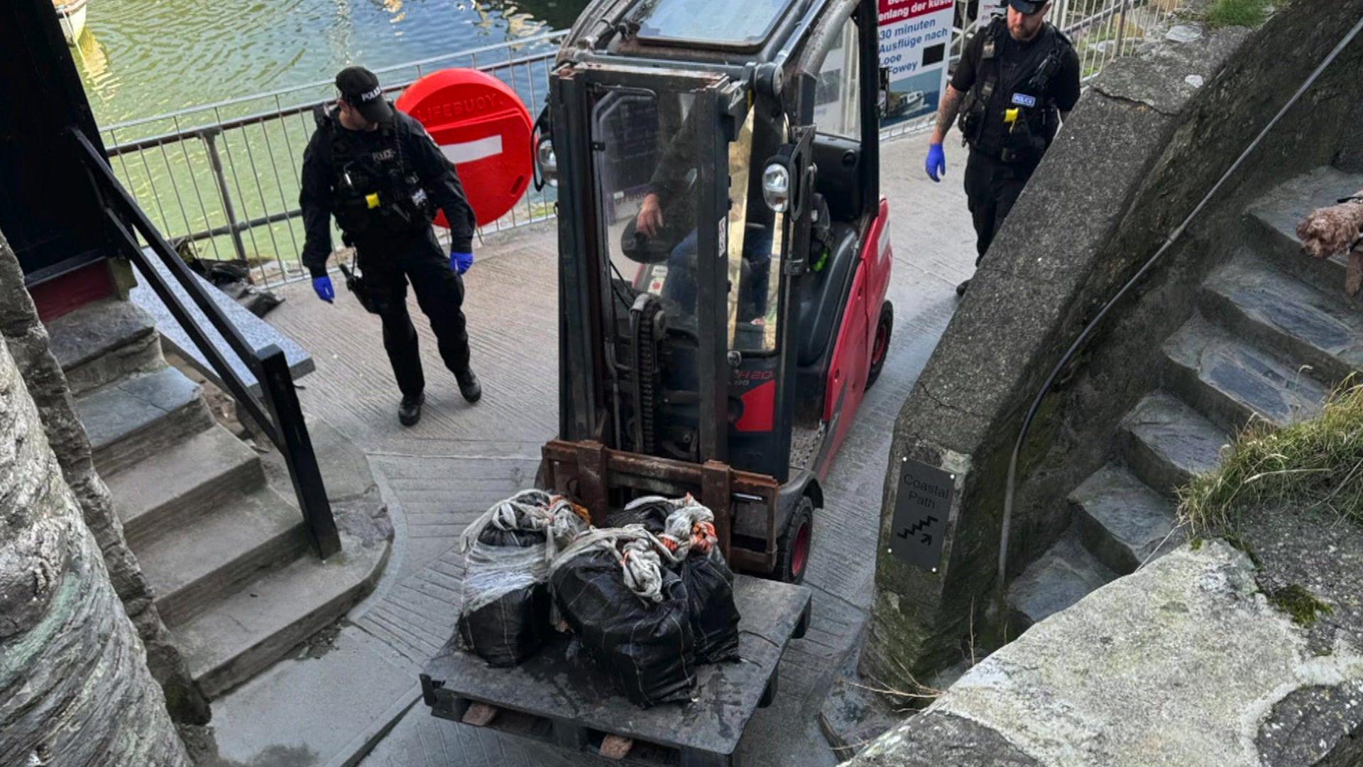 Two police officers either side of a small crane which is holding three bin liners filled with suspected drugs, on either side of the men are two concrete stair paths. Sea is seen in the background.