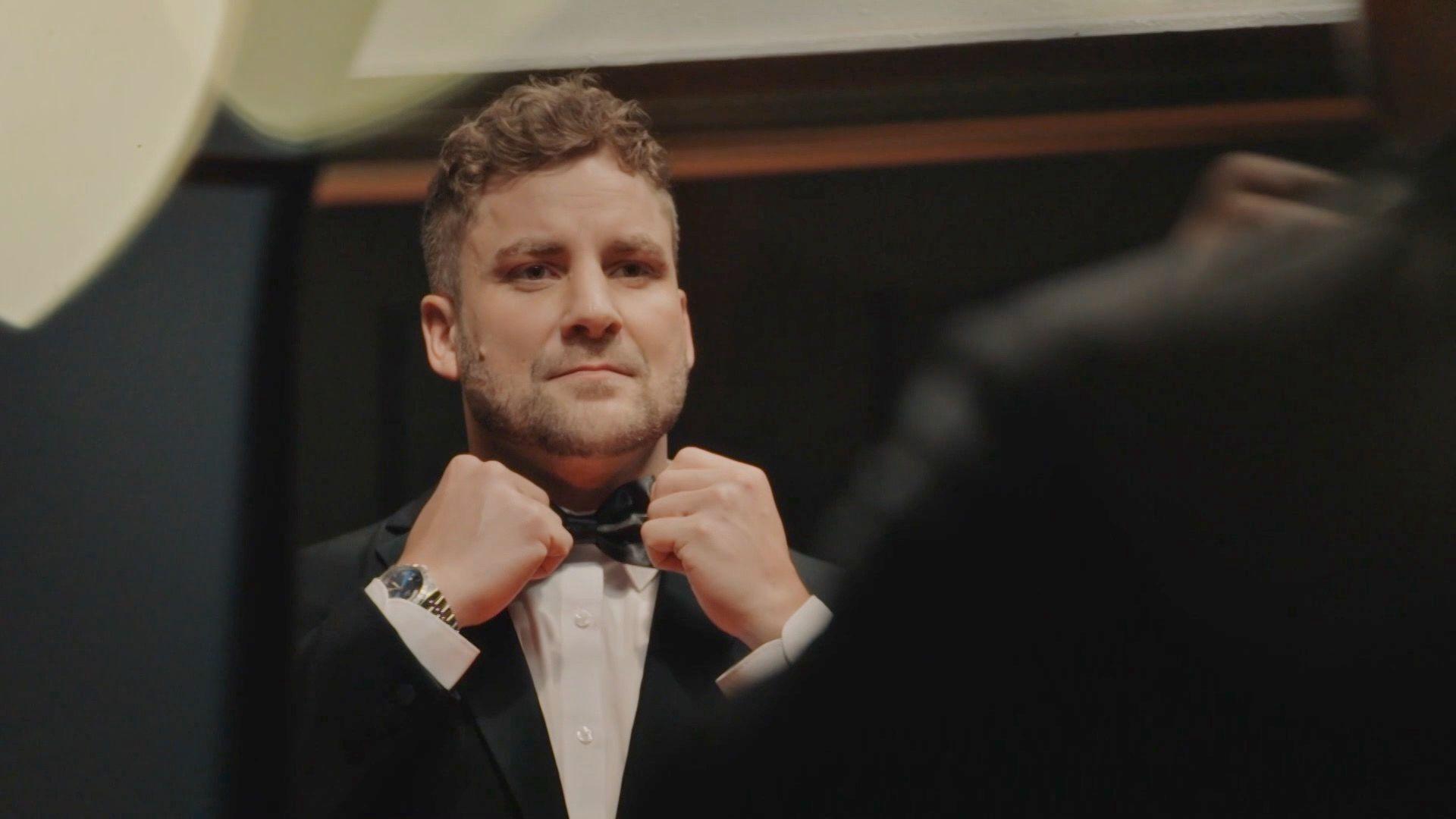 A man looking in the mirror fixing his black bow tie. He has a black suit and white shirt on and short brown hair.