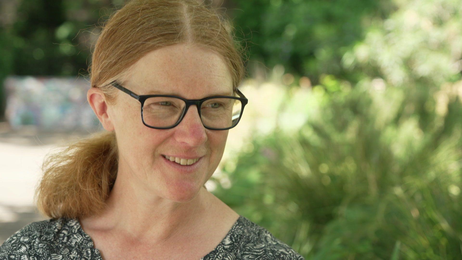 Dr de St Croix is seated on a bench in a garden on a sunny day. She is smiling and has shoulder length hair, which is tied back, and wears glasses. 