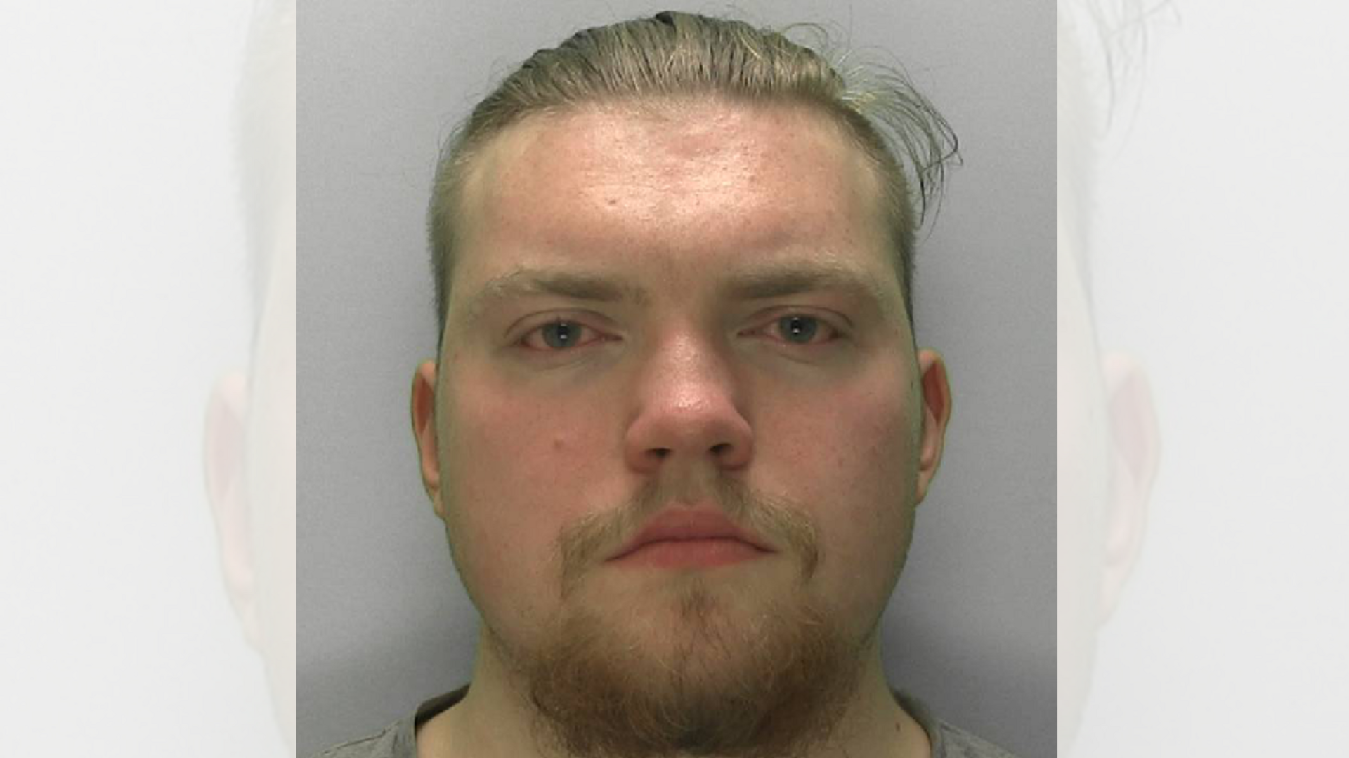 A custody mugshot of a young man staring straight ahead. He has a round face, a light brown beard and moustache and slicked back hair. He appears red in the face.