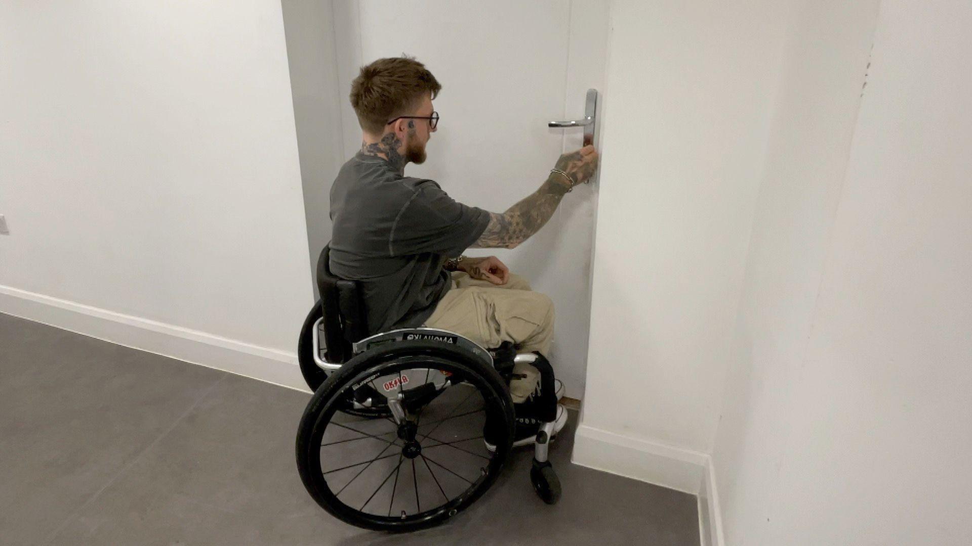Joel Connor-Saunders, wearing a grey T-shirt, using the broken wheelchair in a white room. He is opening the door of the room with a key.