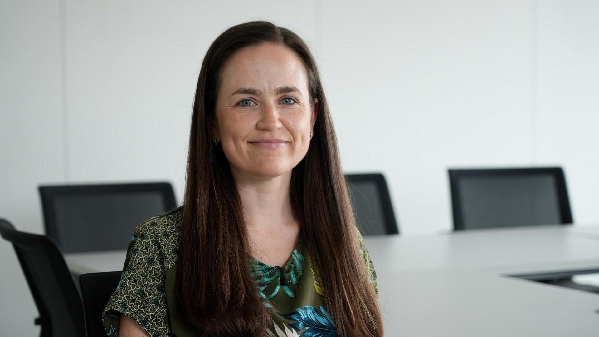 Bethan Evans with long dark hair is sitting in an office with white walls, a desk and black chairs in the background.