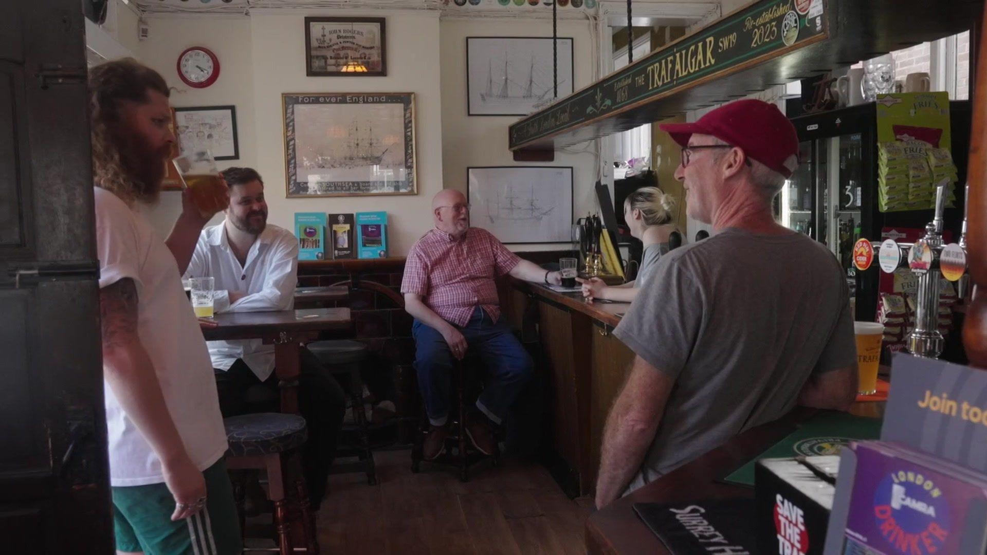 Several people inside a pub talking and drinking, with beer glasses on tables and at the bar, and wall pictures and signs behind them.