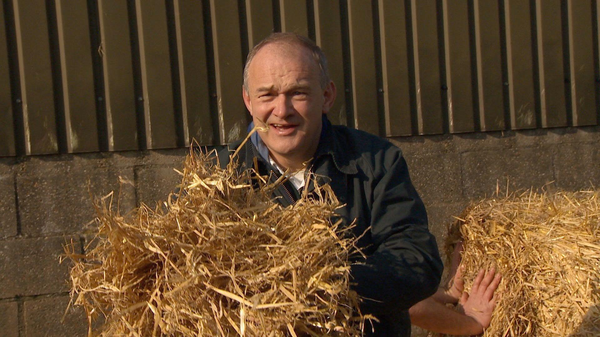 Sir Ed Davey, wearing a dark jacket, clutches a pile of hay.