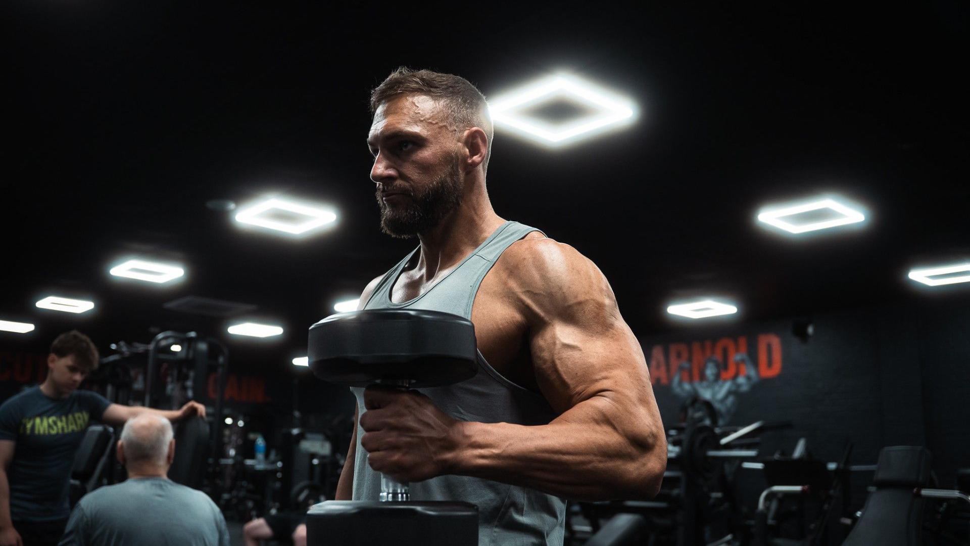 Brendan Pearl is holding two 25kg dumb bells, behind him are other people on various machines and equipment in a gym. He has a serious expression as he tenses his arm to lift the weight. 