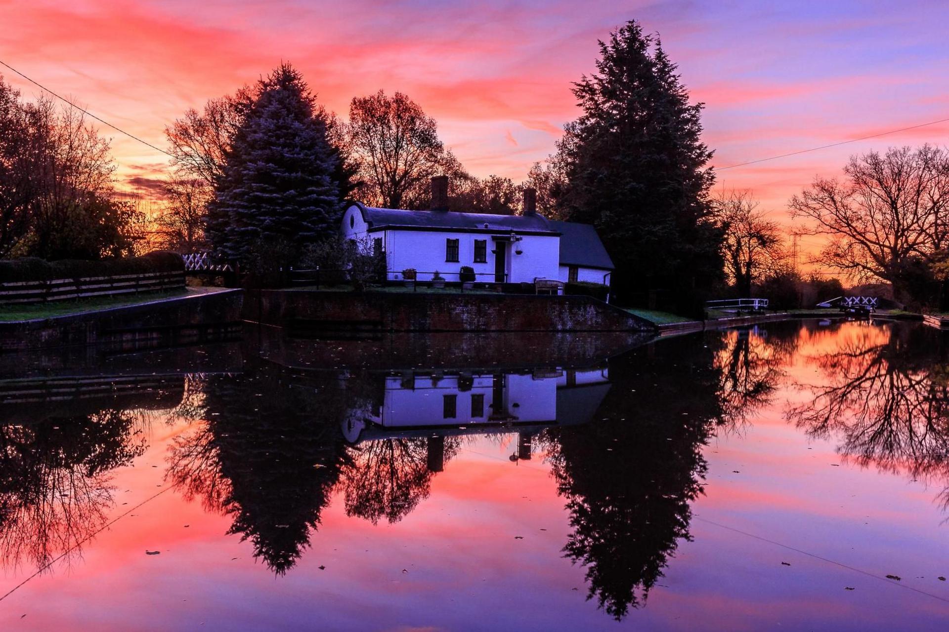 The reflections of three large trees and a white building are seen in the water. The bottom half of the photo is the water, which also includes the sunrise reflection