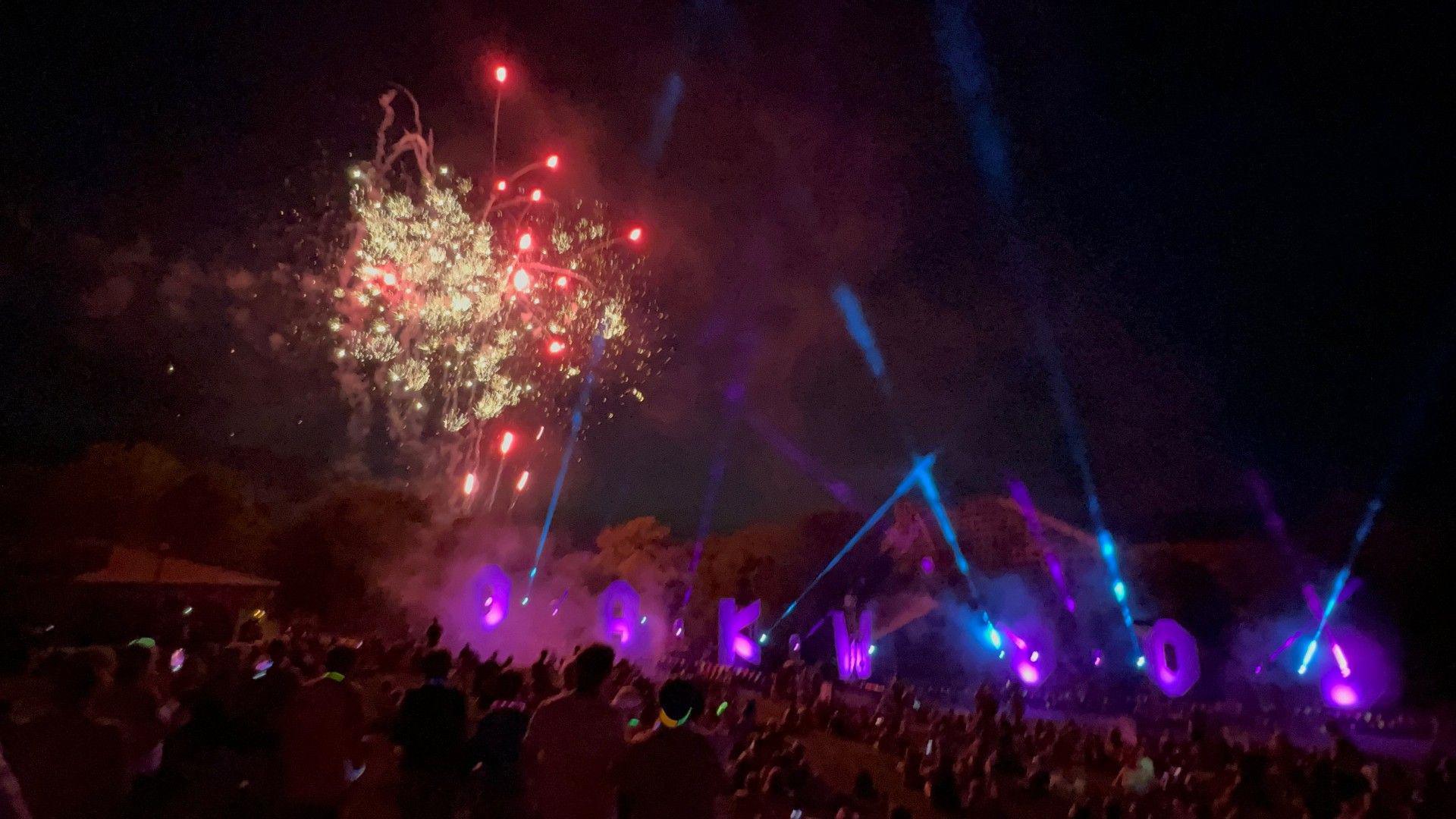 Strobe lights can be seen emanating from spots around the Oakwood sign. They are blue and purple in colour. A firework display can be seen going off in the background with yellow and red colours in the sky. A big group of people can be seen in the foreground of the image looking at the display.