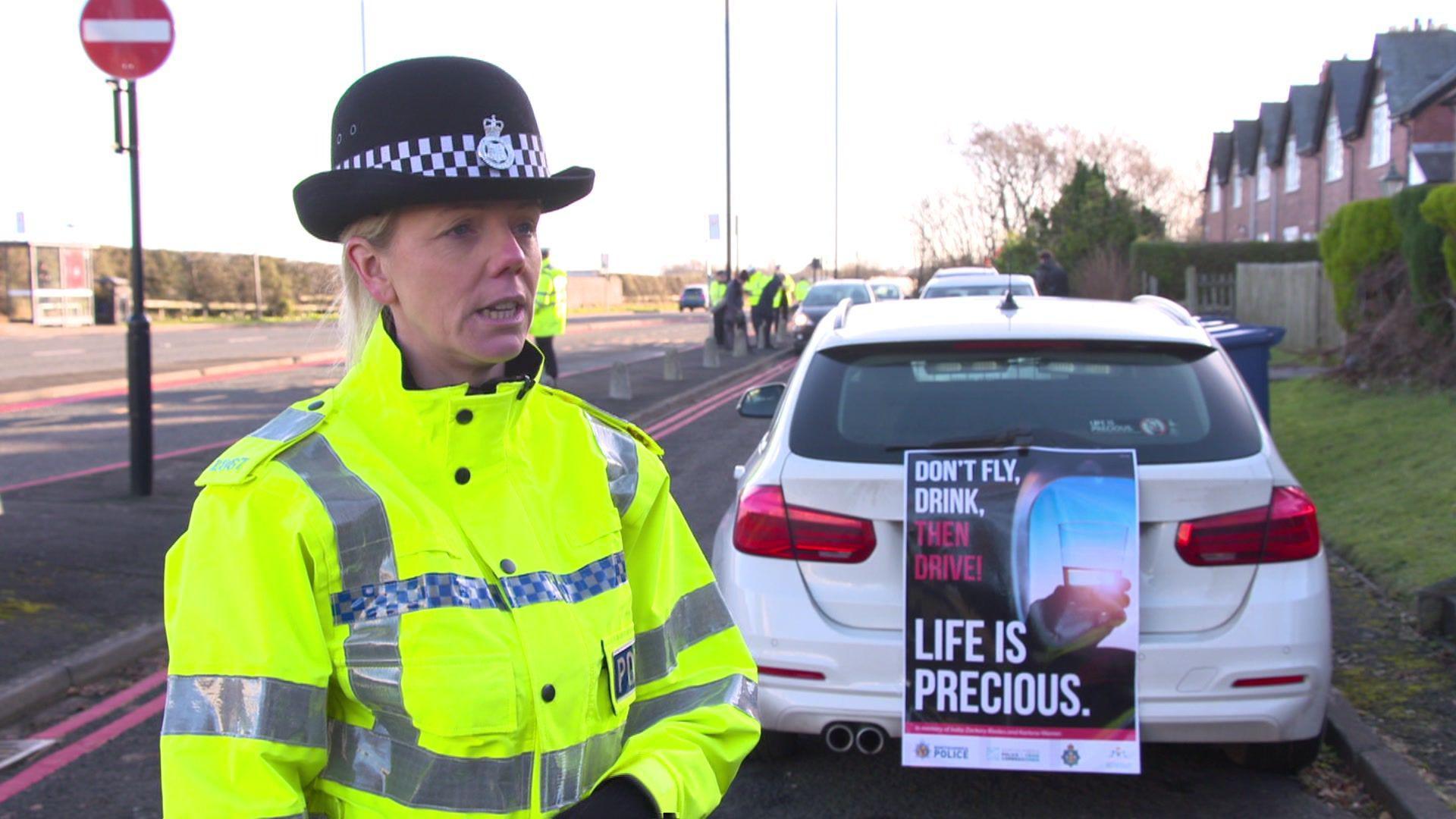 Det Con Natalie Horner. She is wearing her police hat and high-vis jacket and is standing next to the road where the police were stopping drivers. A police car is parked behind her with a poster which reads, 'don't fly, drink, then drive!'