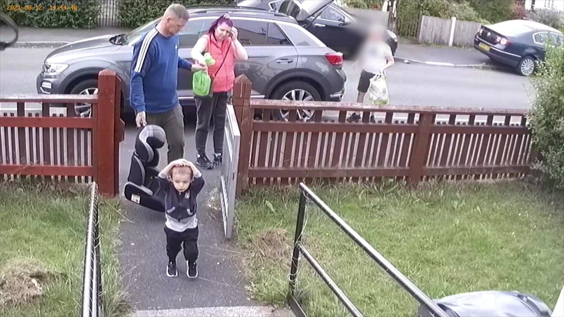 Llun o Ethan yn cerdded tuag at y tŷ gyda'i freichiau ar ei ben. Mae ei dadcu a'i mamgu yn cerdded tu ôl iddo