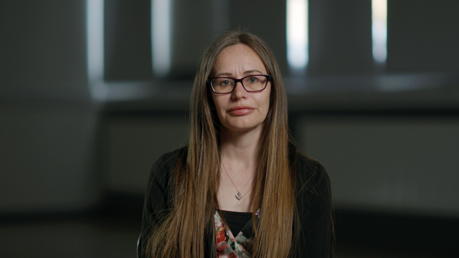 A photo of Susan Christie. She is looking at the camera against a dark internal background. She has long brown hair and glasses.