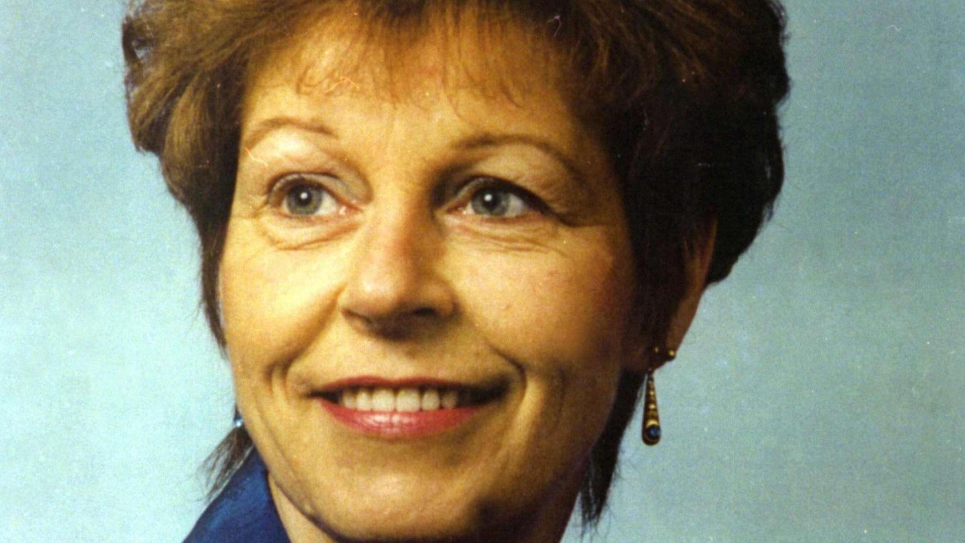 Sandie Bowen is pictured in a portrait looking to the side and smiling, wearing earrings and a blue dress. She has short bouffant brown hair.