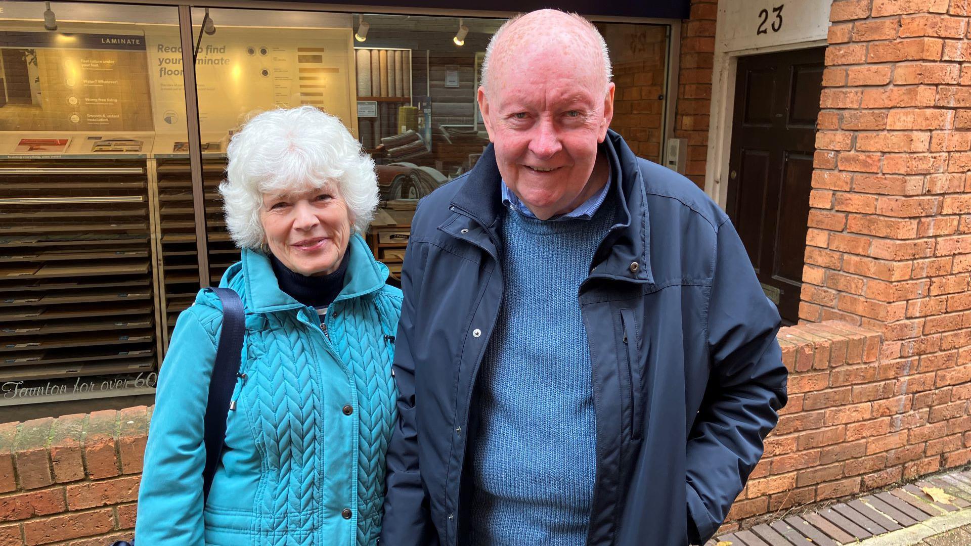 Jackie and John stood side by side - Jackie is in a blue coat, while John is in a blue jumper and blue coat. Both are looking at the camera.