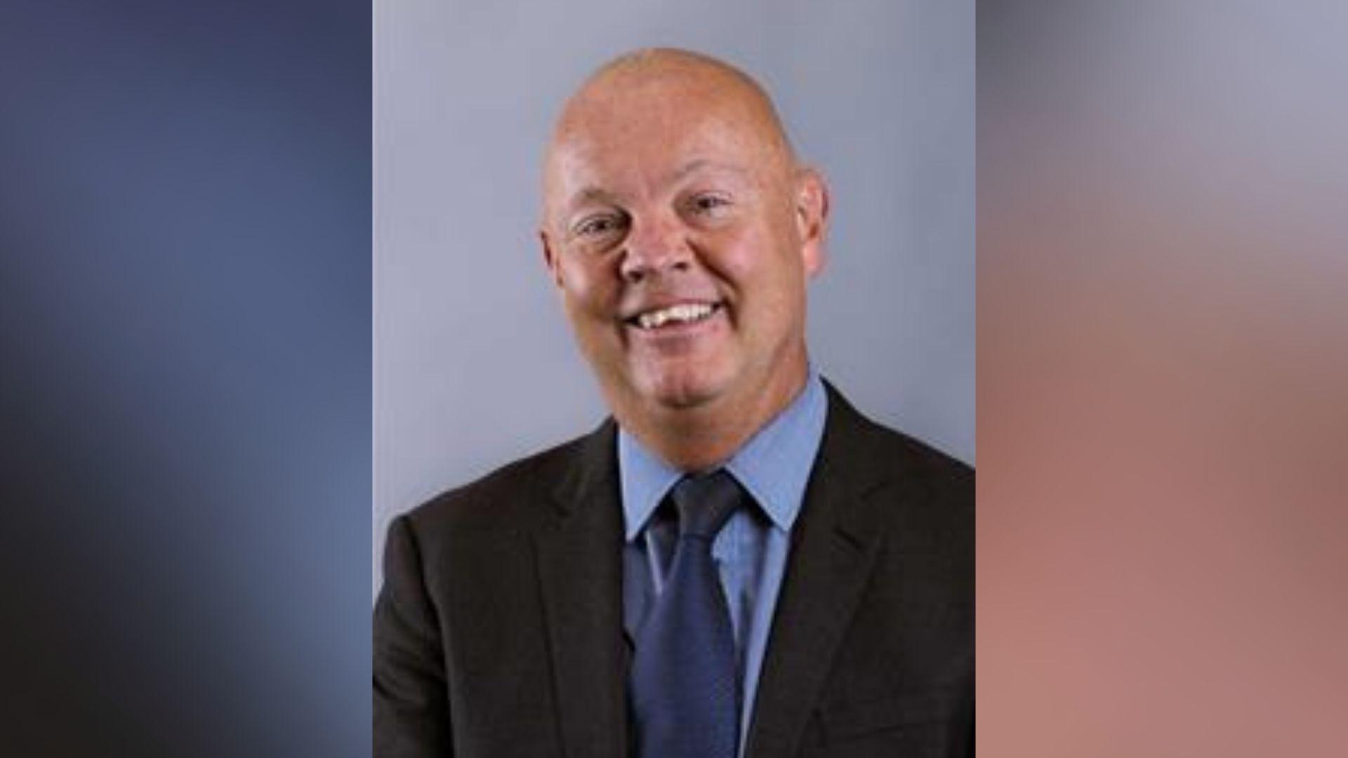 A head and shoulders shot of a smiling bald man in a suit and tie on top of a blurred background