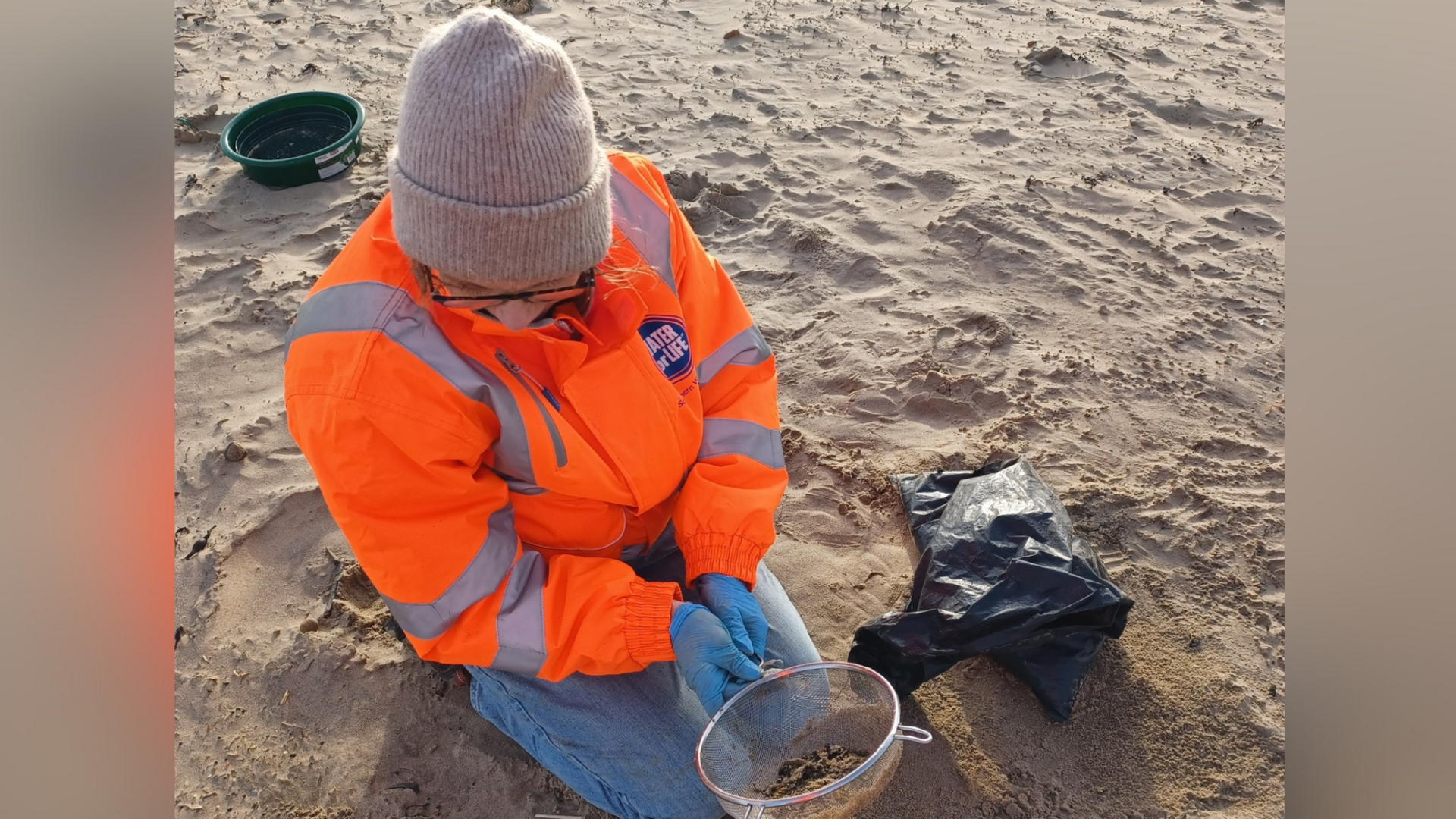 A Southern Water employee on Camber Sands in East Sussex cleaning up plastic pellets. The employee is wearing jeans and an orange high-viz coat. She is holding a utensil full of pellets.