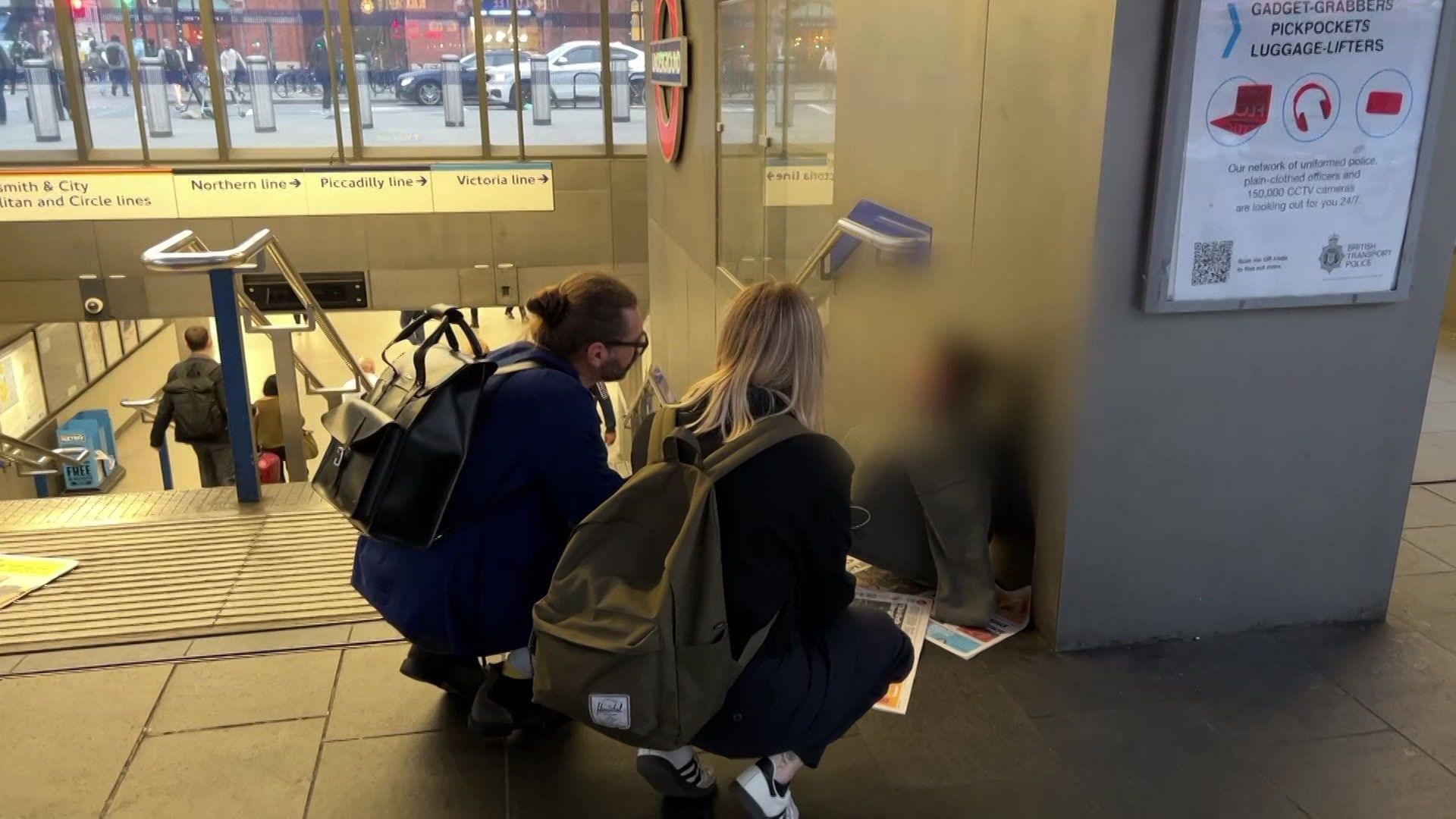 A male with a ponytail and classes and a female with shoulder-length blond hair crouch down to speak to a person sitting on some newspaper at the top of some stairs.
