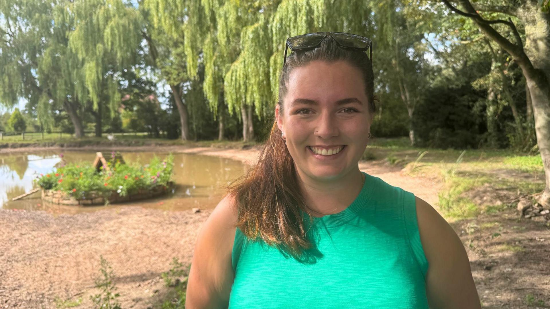 A woman in a green vest smiles next to a shallow pond, surrounded by trees.