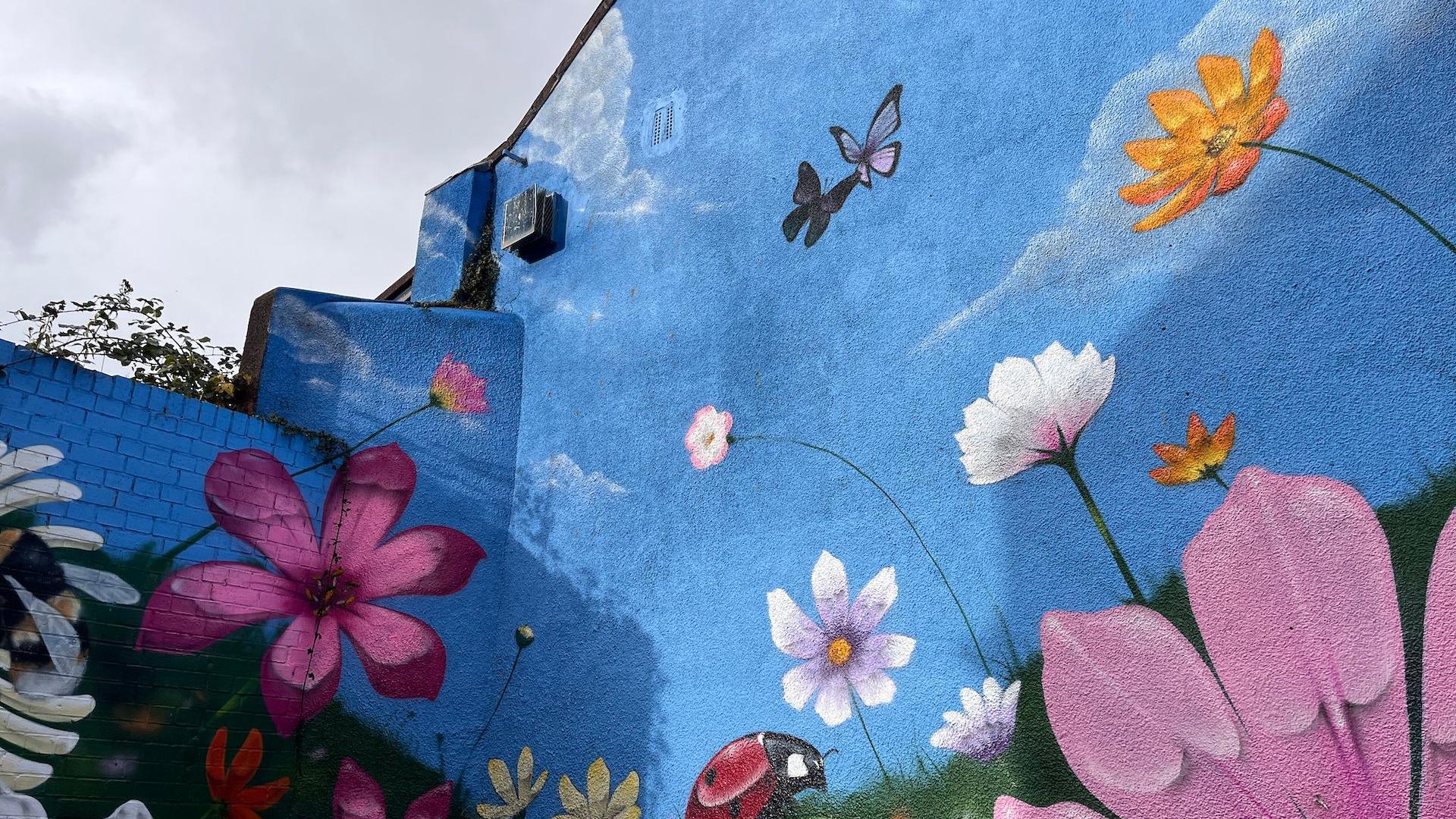 A blue painted wall, with pink, yellow, white, and purple flowers on, with butterflies on it. The sky in the corner is grey.
