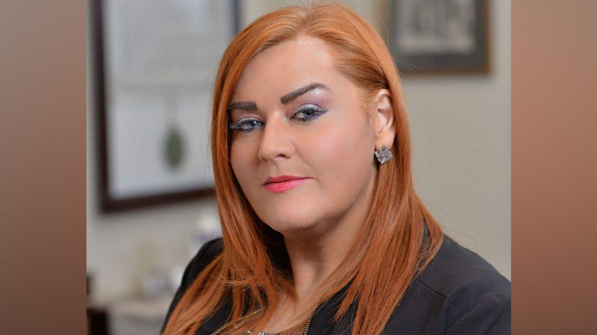 A woman with red hair looks at the camera. She wears pink lipstick and has a black blazer on. She has a silver diamond earring visible in her left ear as her hair is tucked behind.