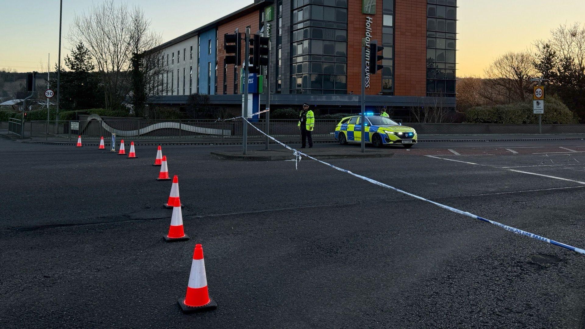 A road closed with cones across the road, police tape and an officer with a car.