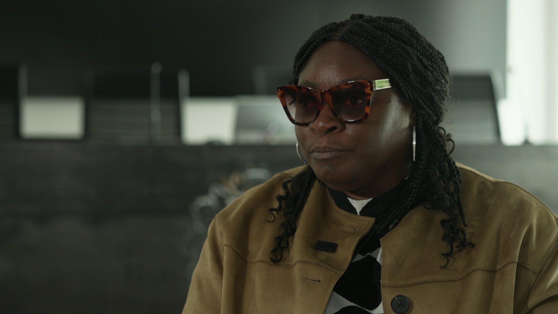She is sat inside, wearing a camel coloured coat and a black and white top. Shehas silver hoops in and wears tortoise-shell sunglasses. She is looking to the side of camera, and has medium-length black hair. 