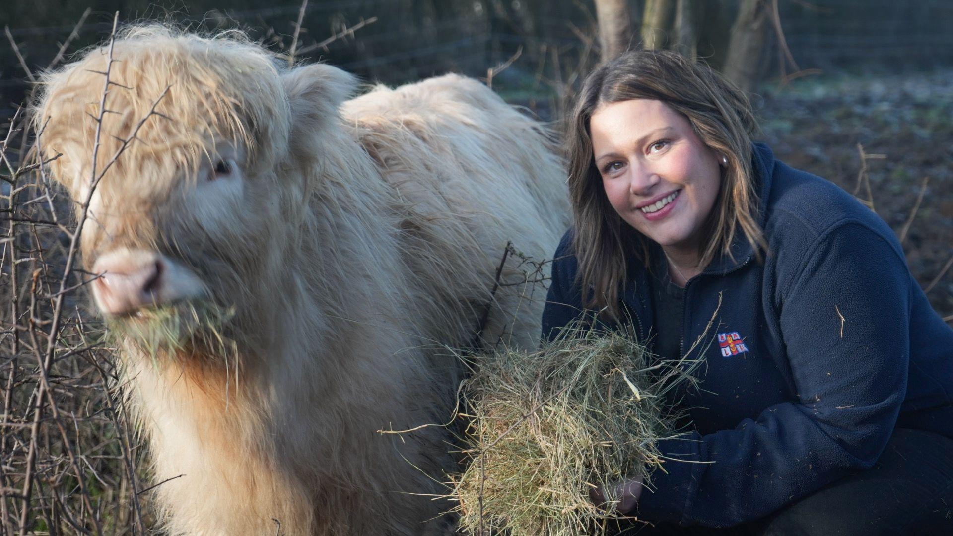 Laura Chimes, crouching down next to a Highland cattle. Laura is wearing a navy blue fleece with an RNLI logo on the chest. Laura is holding a handful of hay which she is feeding to the cow.