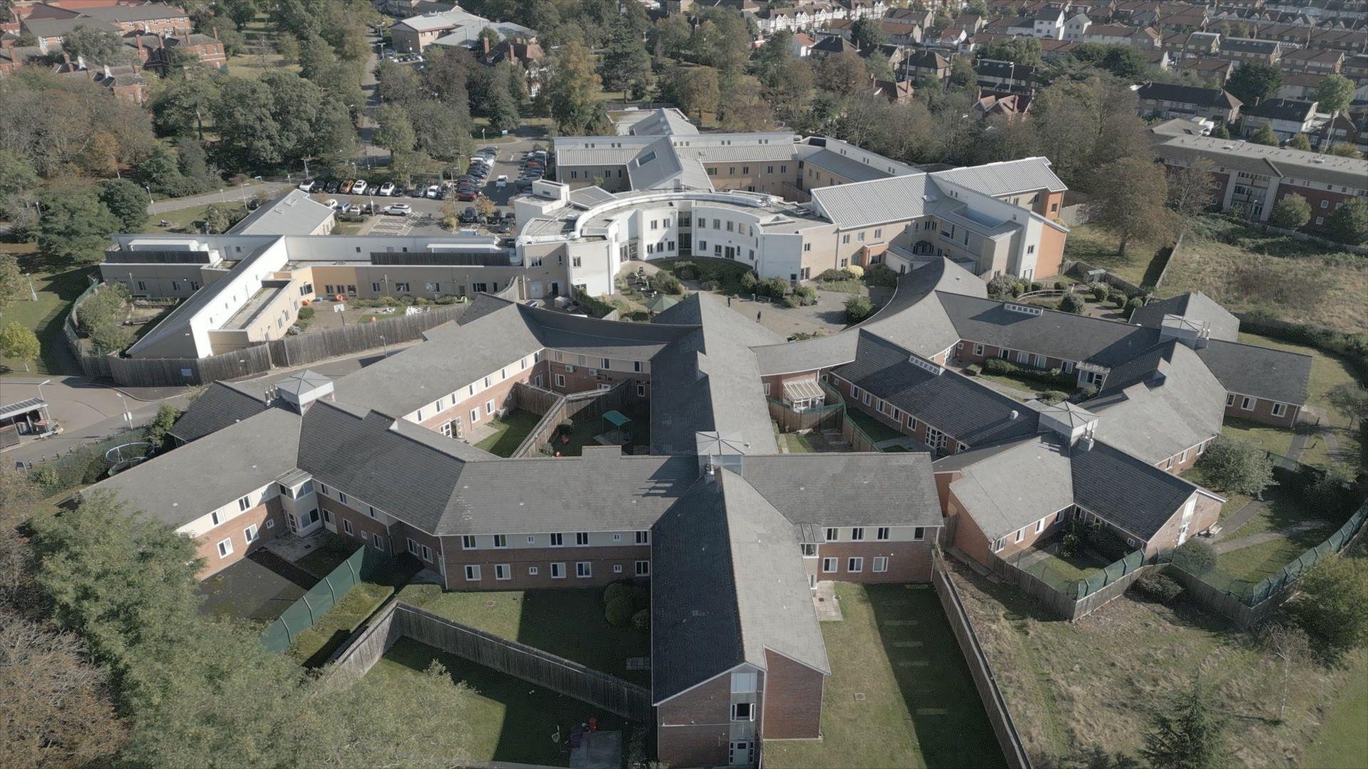 An aerial photograph shows Goodmayes Hospital from above. Buildings span out from a central circular area. It is surrounded by grass and trees.