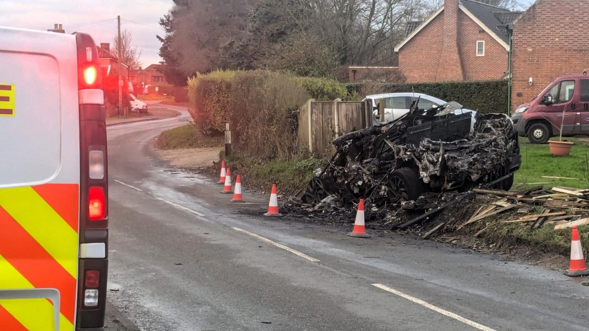 A police van with its lights on parked on the opposite side of the road to the burnt-out car. A line of road cones have been placed around the wreckage. 