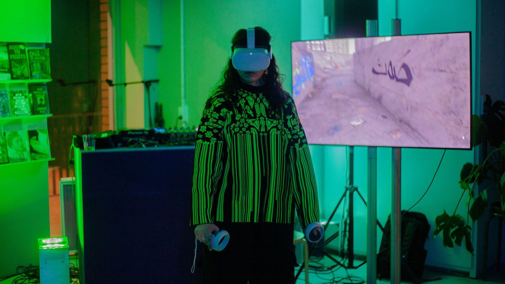 A person with long curly black hair wearing a green and black vertical stripy shirt. The person is holding two white controllers in their hands and is wearing a white VR headset on their head. They are stood in front of a TV screen in a green room