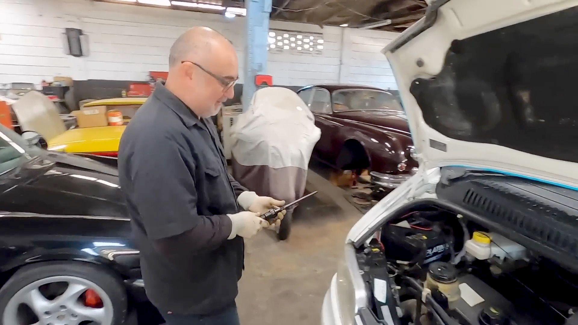 A male mechanic in blue overalls stands holding a tool and looking into the open bonnet of the white camper van, behind him other cars are visible