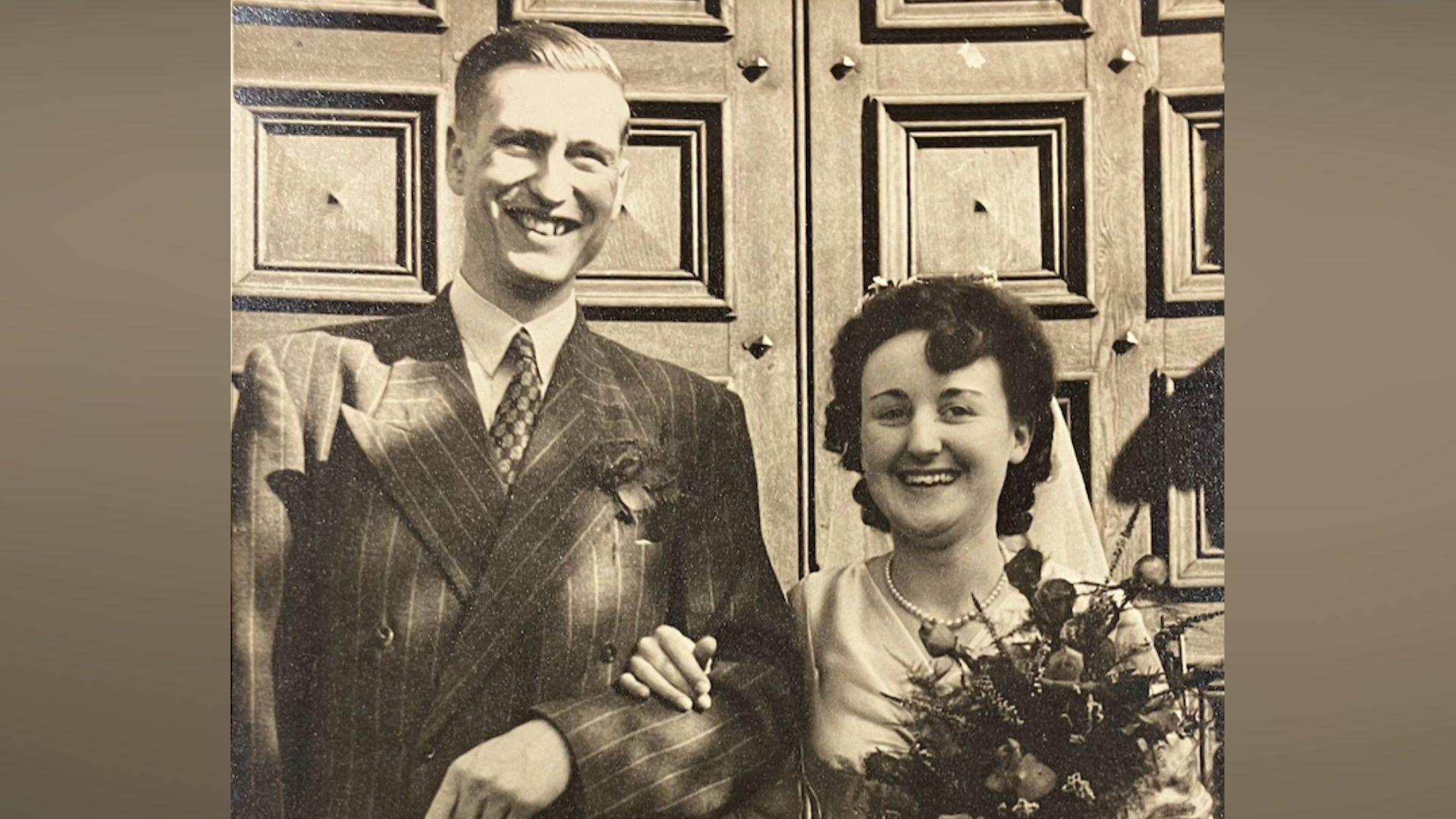A black and white image of a young man and woman on their wedding day. They stand in front of a large wooden door and smile at the camera. The man has short hair and wears a suit, tie and shirt. The woman wears a white wedding dress and holds a bouquet of flowers in one hand while her other hand rests on the man's forearm. 