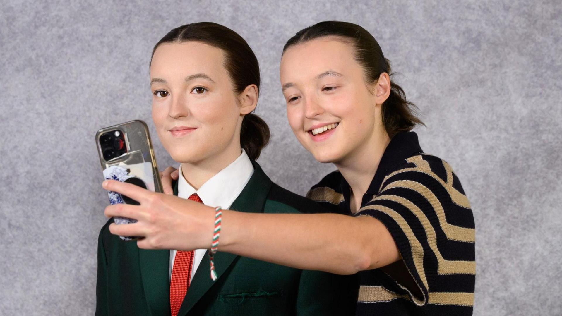 Bella Ramsey wearing a black and cream hooped top stood behind a waxwork of them. They have a phone in their hand and are taking a selfie with the figure.