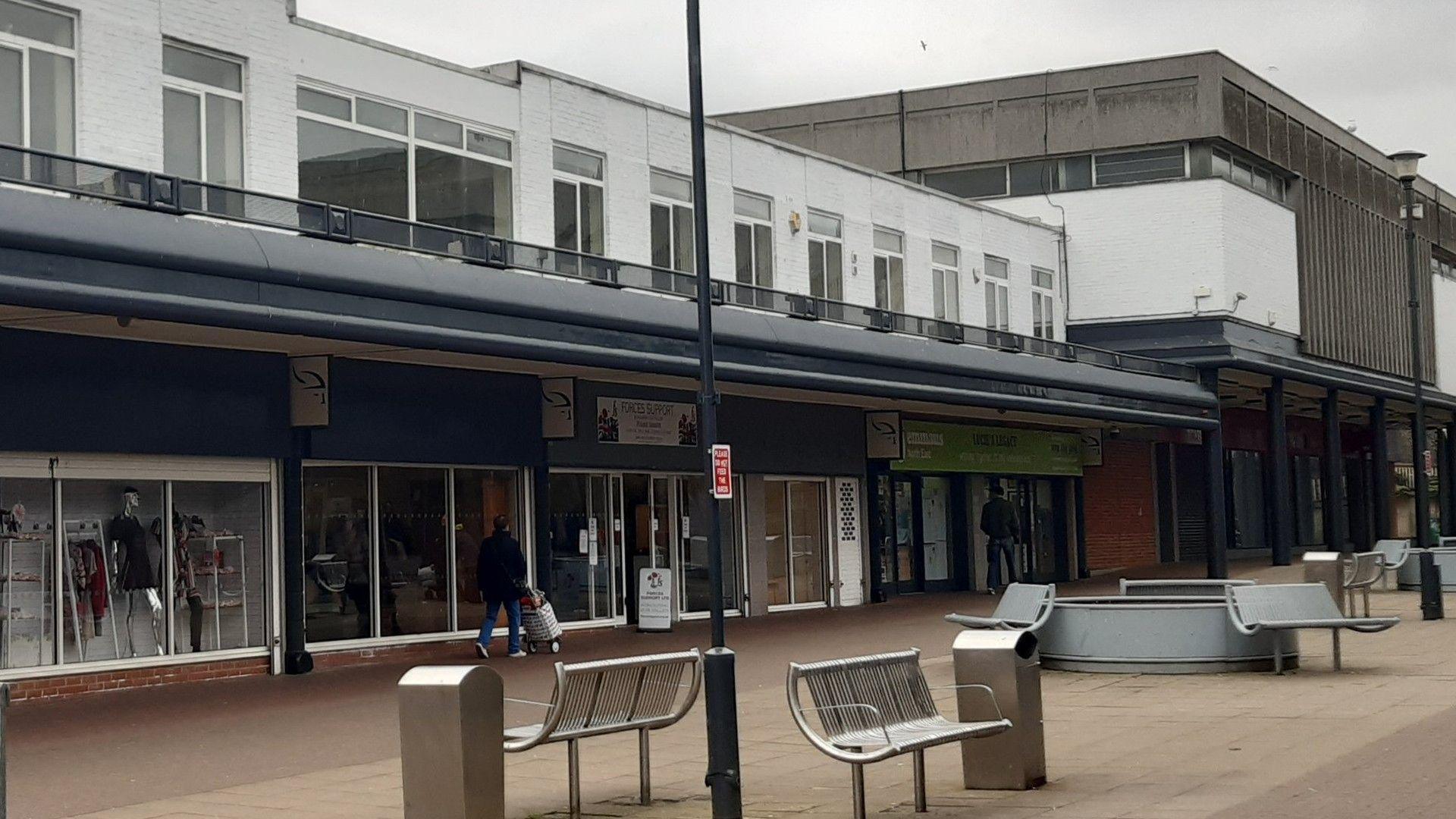 A row of retail units which are housed in a white 80s-style building. The high street has silver benches and bins outside.
