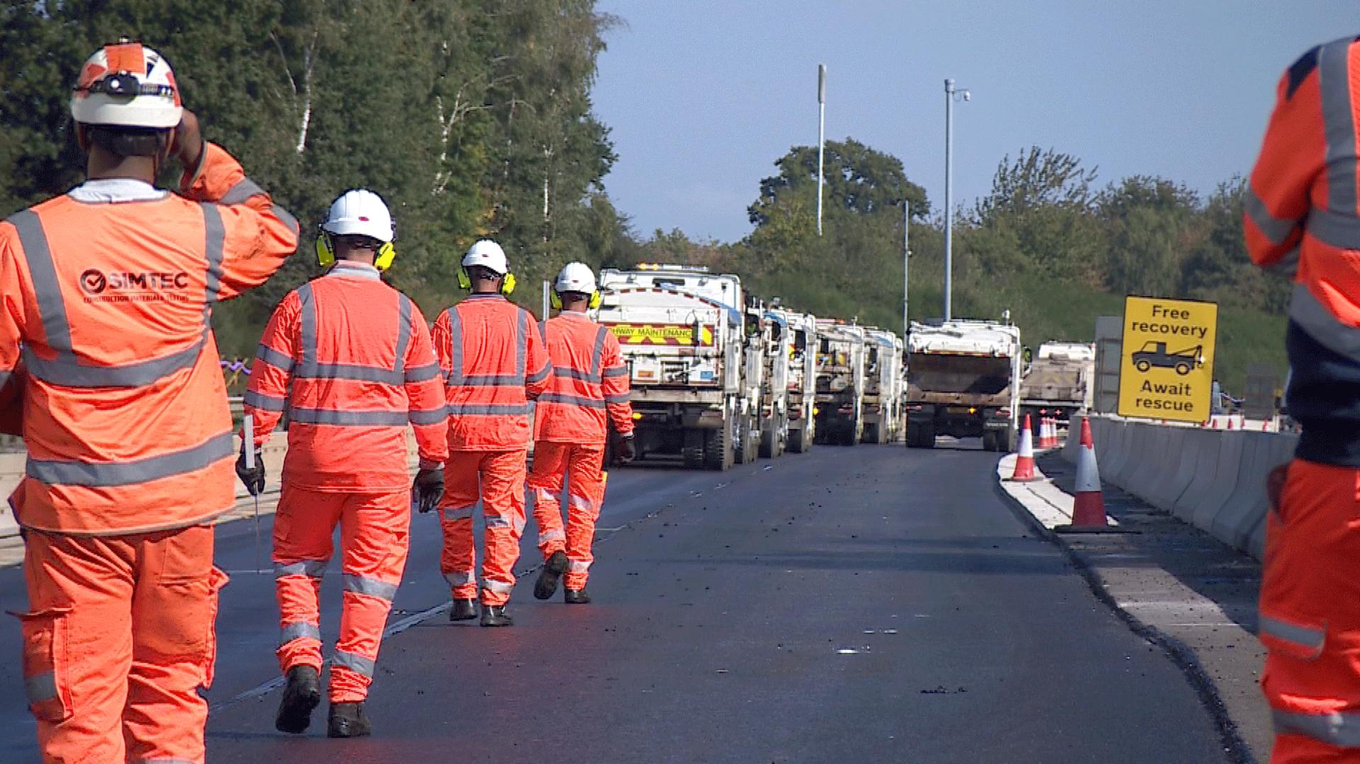 A line of workers in orange uniforms and hard hats march up the newly-surfaced motorway towards a row of tarmac trucks.