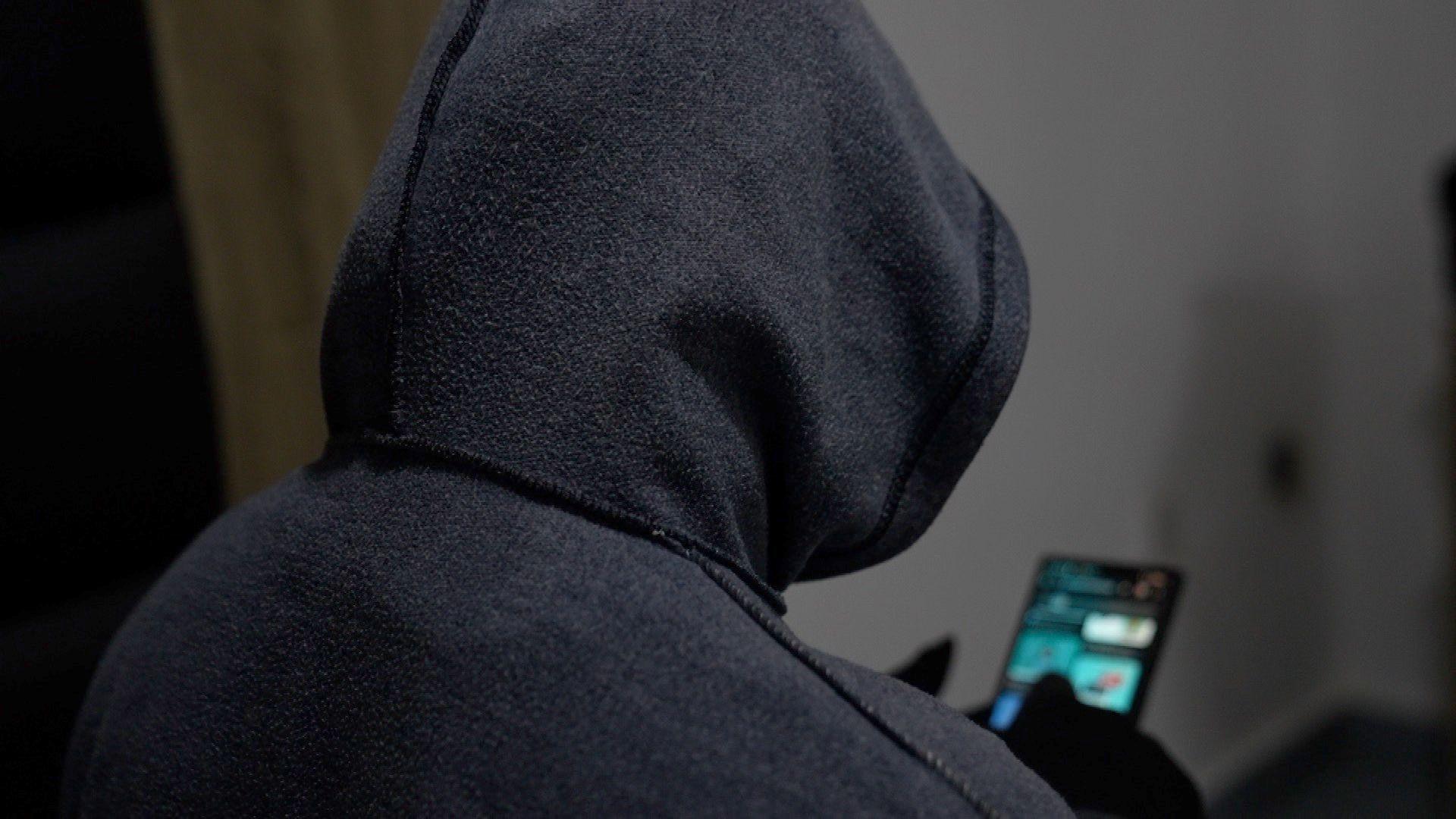 A picture from behind of a hooded person viewing a mobile phone. The person's identity is hidden.