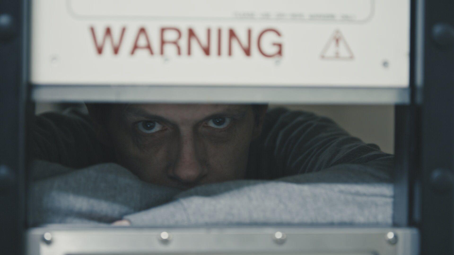 Man staring through slot in door with word "warning" above in red