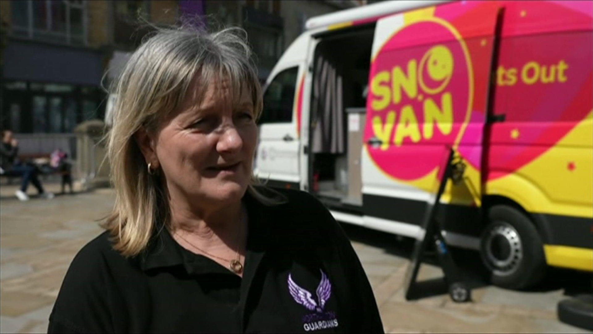 A woman with blonde hair in a black polo with angel wings on it stands by a white van with SNO or "Safer Nights Out" written on its side in a yellow font on a pink background. 