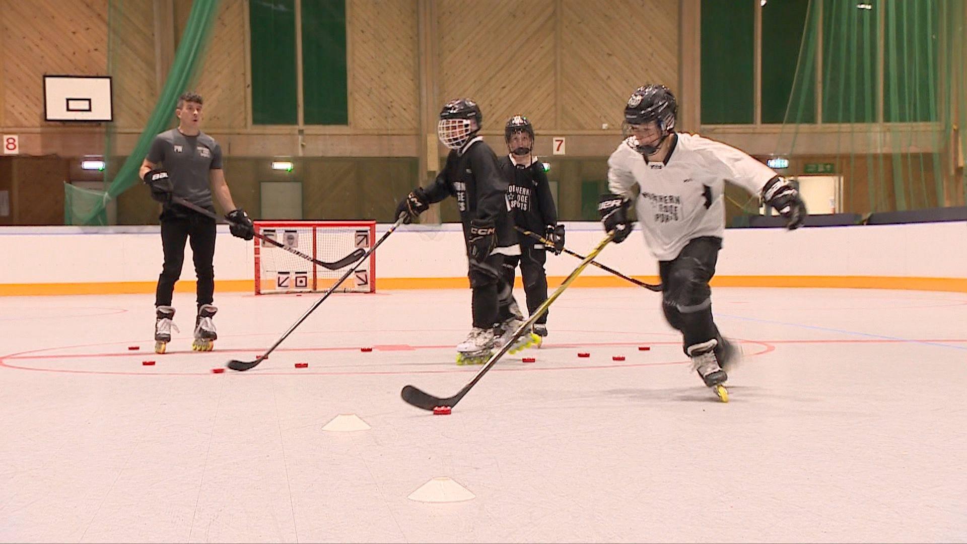 Four people playing roller hockey in a big hall. They are holding large hockey sticks and wearing oversized black and white jerseys and helmets with face guards.
