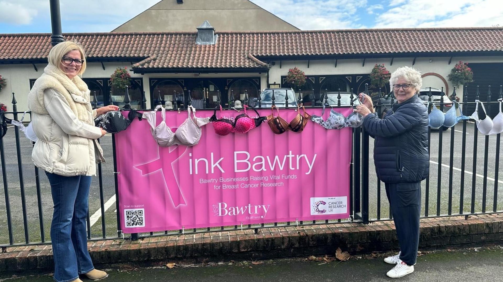 Two women are hanging bras on the fence outside a business in Bawtry. A banner attached to the fence reads: 'Pink Bawtry. Bawtry Businesses Raising Vital Funds for Breast Cancer Research.'