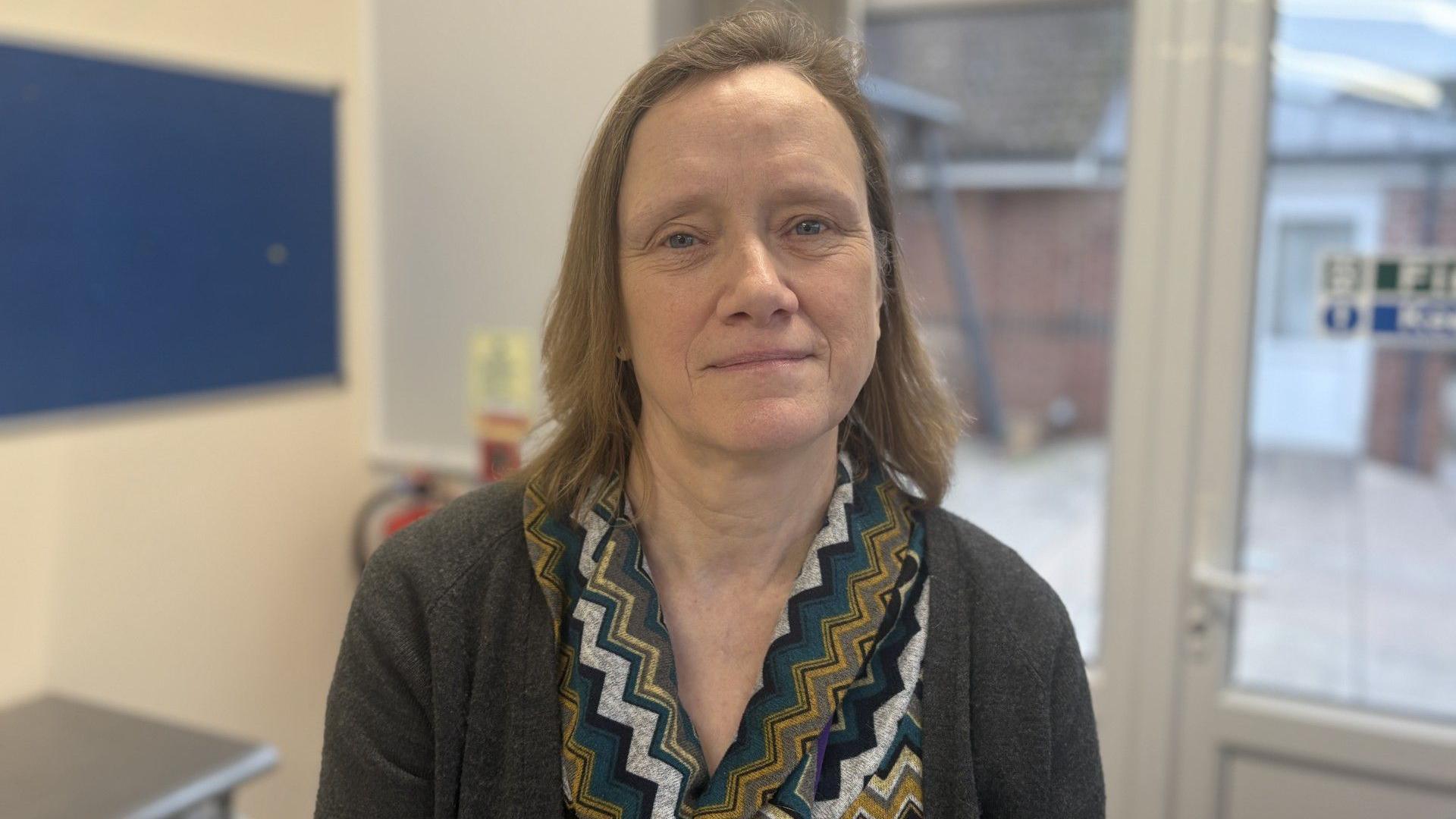 A woman with light brown, shoulder-length hair is sitting in a medical setting. She is wearing a grey cardigan and zig-zag patterned top.