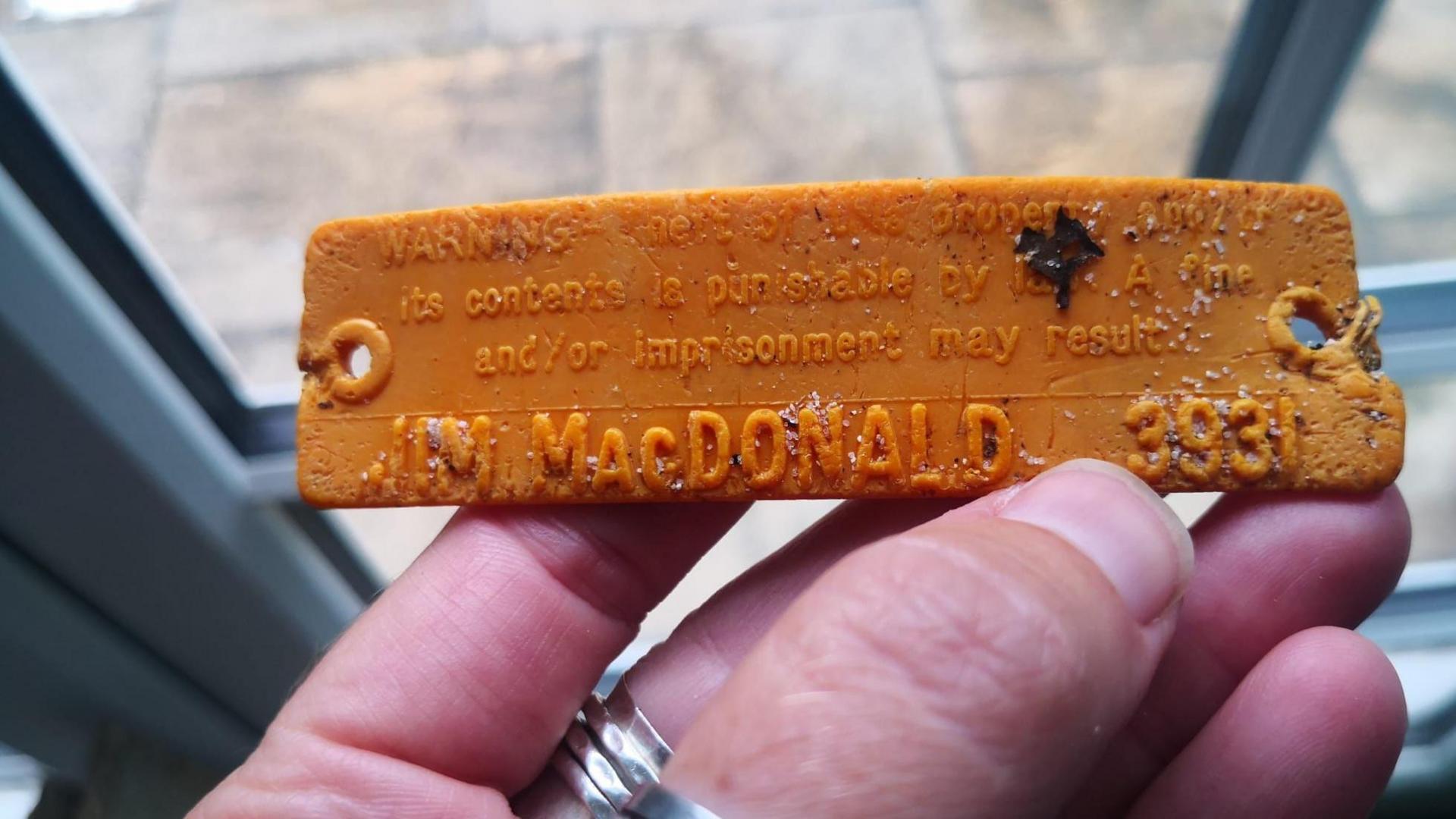 An orange tag with the name 'Jim MacDonald' on it. Nikki is holding it in her hand in front of a window. 