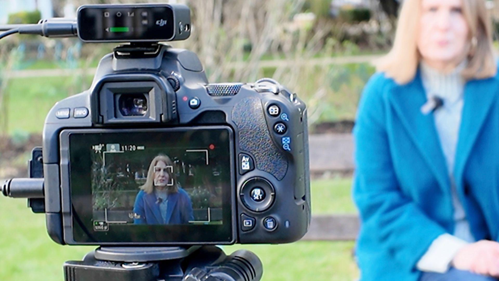 The back of a camera pointing at Julie Gould. She can be seen in the distance in a blue coat and sitting on a park bench.