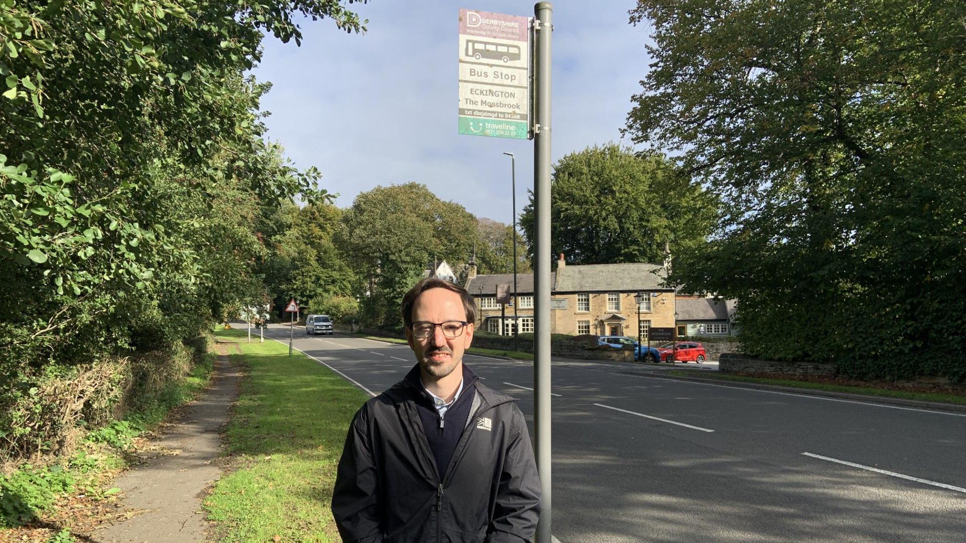 A man with short black hair is wearing glasses and a black raincoat. He is stood by a main road in front of a bus stop.