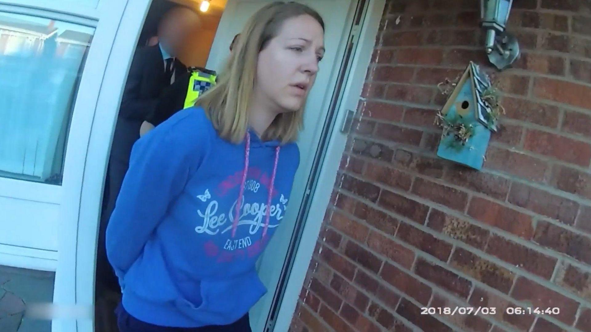 Police bodycam footage showing Letby, wearing a blue hoodie, being led from her the front door of a house with her hands behind her back. 