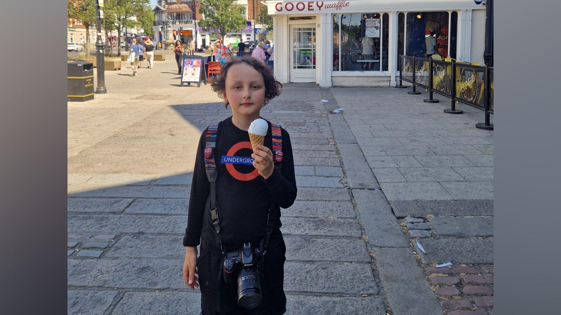 A picture of George stood outside a 'Gooey Waffle' shop in London on the day of the challenge. George is wearing all black, and his top has a TfL roundel on it. He has longish brown hair and is holding an ice cream. He also has a bag on his back and a camera around his neck.