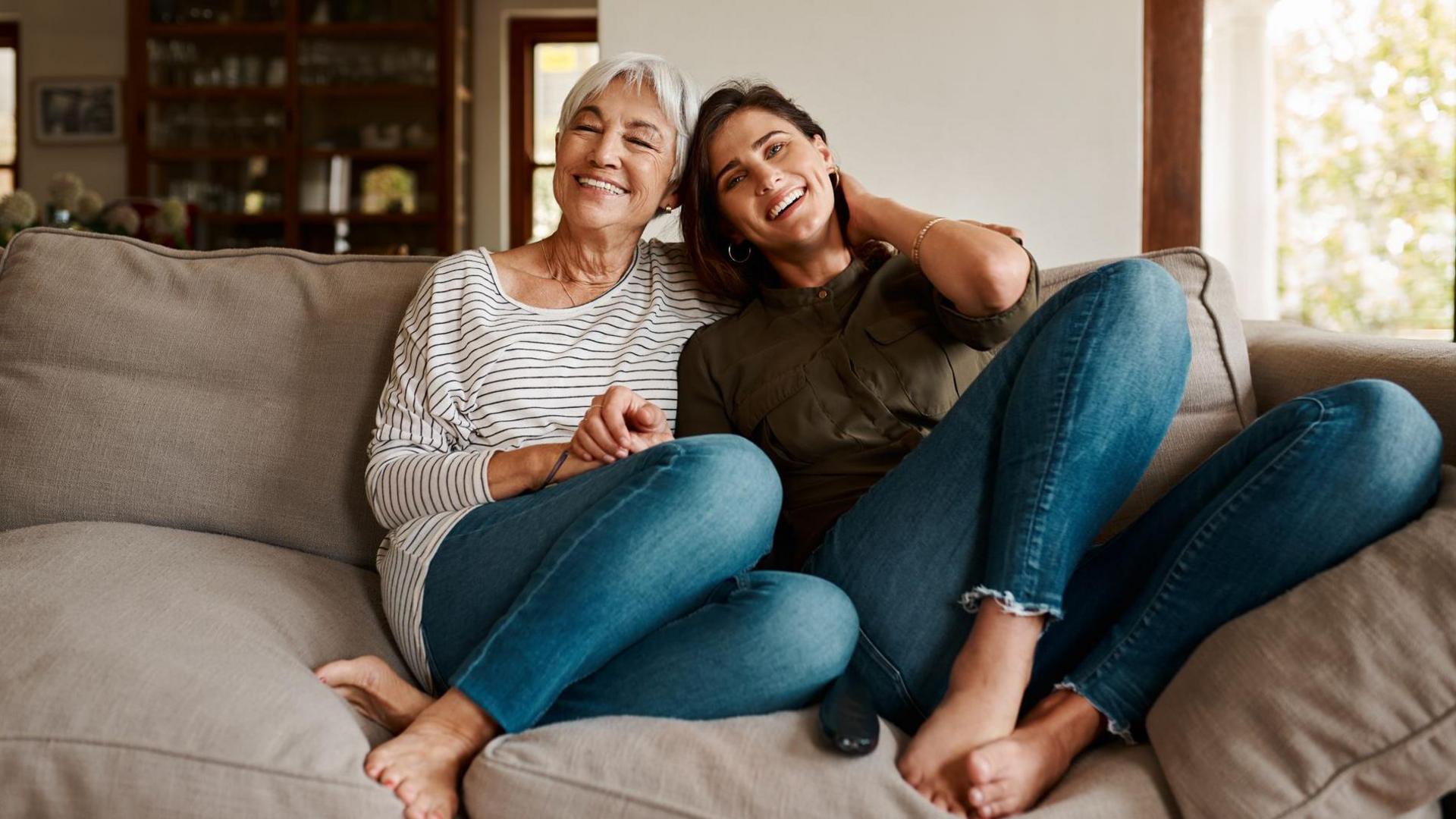 Two generations of women sit on a sofa smiling and holding hands with a cabinet and picture in the background.