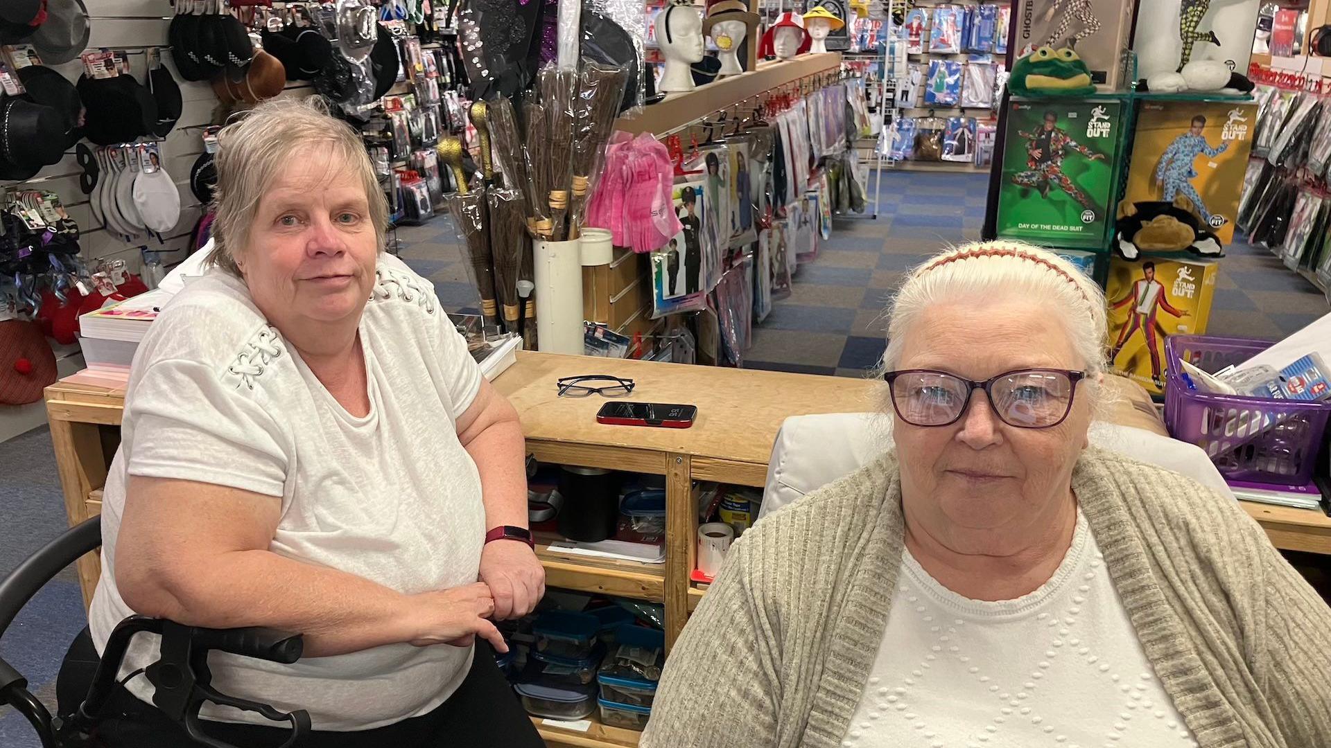 Mandy is on the left and she is wearing a white tshirt, and is sat on a mobility aid in front of a desk. Next to her is Dorothy, wearing a white top and beige cardigan. Behind them is the interior of the fancy dress shop.