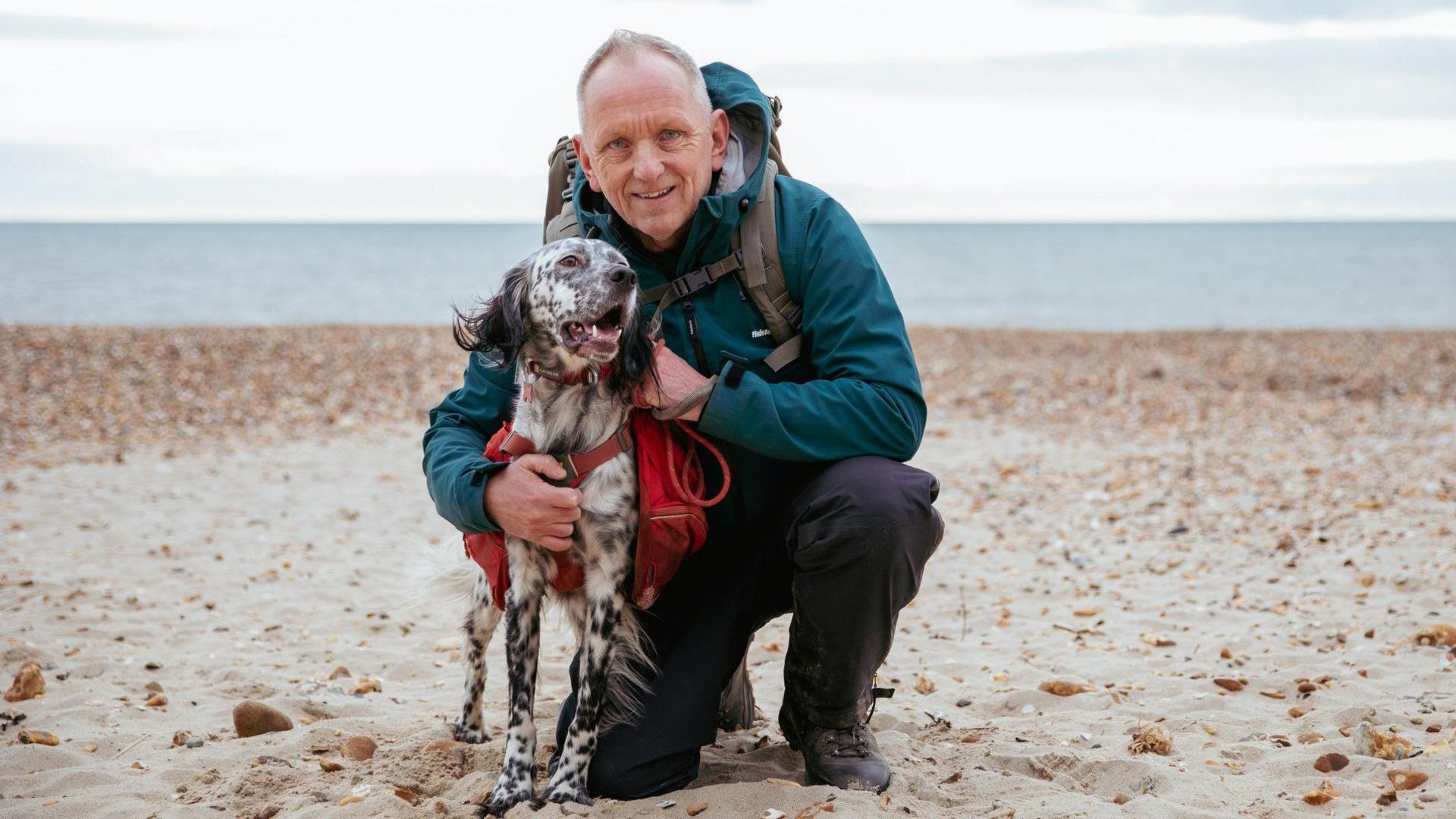 Louis and Nick pictured on a sandy beach, with grey sea visible in the background. Louis has white fur with black markings and black fluffy ears, and wears a red harness and bag. Nick is crouching next to him, wearing hiking books, dark trousers and a dark teal raincoat. 