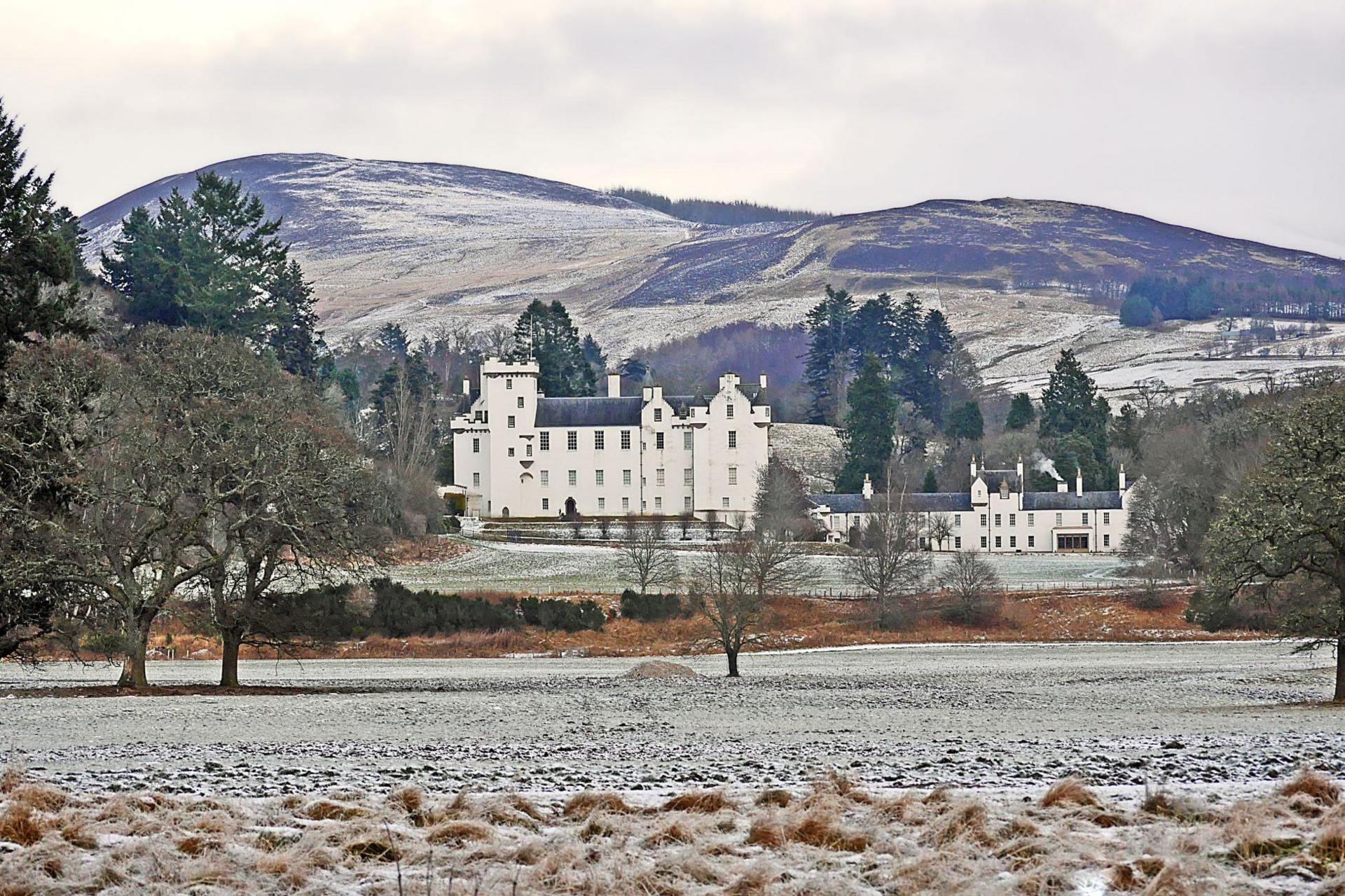 The white-walled castle is pictured on a chilly day. There is frost and a dusting of snow on parkland around the castle. The parkland is dotted with trees and large hill rises behind the castle.