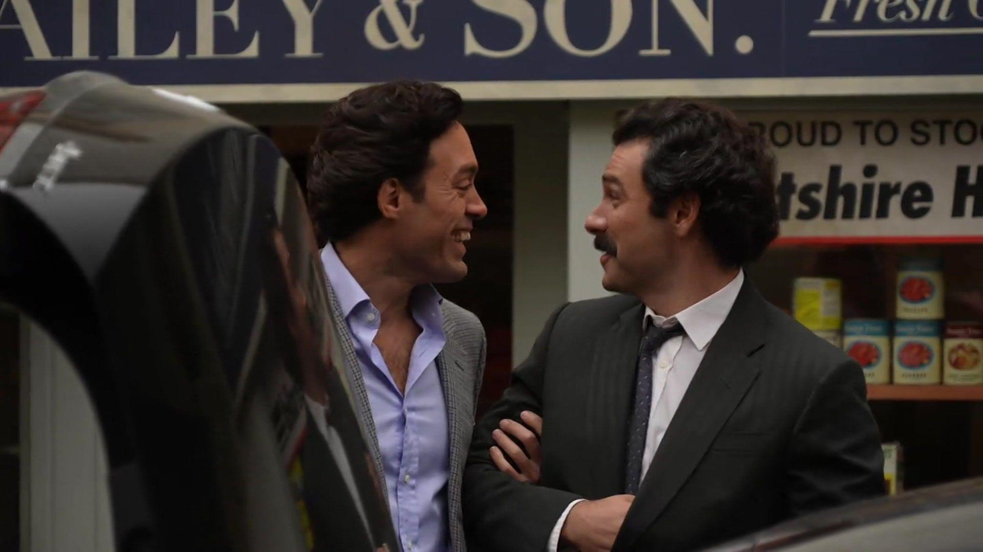 A candid shot of Alex Hassell and Aiden Turner talking to each other on set before filming. Both are dressed smartly in suit jackets, with a car boot up in front of them and a shop behind them